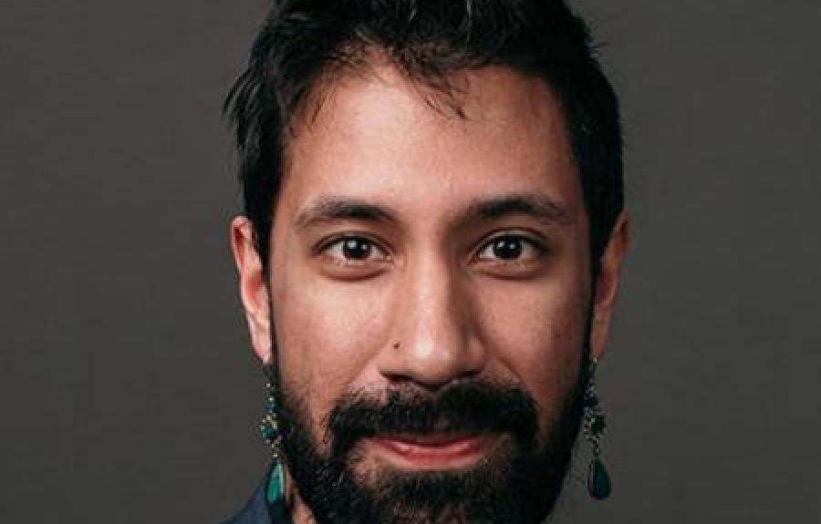 Person wearing a dark, buttoned shirt with a subtle texture, accessorized with a thin necklace and a dangling earrings. The background is a plain, dark gray studio backdrop.