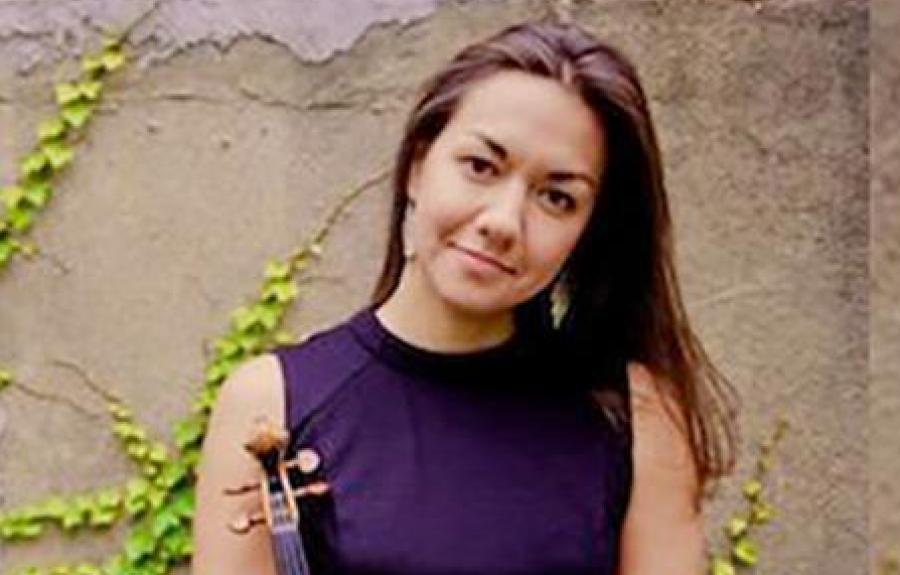 A person in a sleeveless dress stands outdoors holding a violin upright in front of them. They are positioned against a textured wall with green ivy trailing across it. The violin is centered, and the setting has a calm, natural atmosphere.