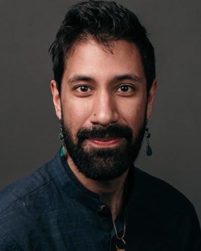 Person wearing a dark, buttoned shirt with a subtle texture, accessorized with a thin necklace and a dangling earrings. The background is a plain, dark gray studio backdrop.