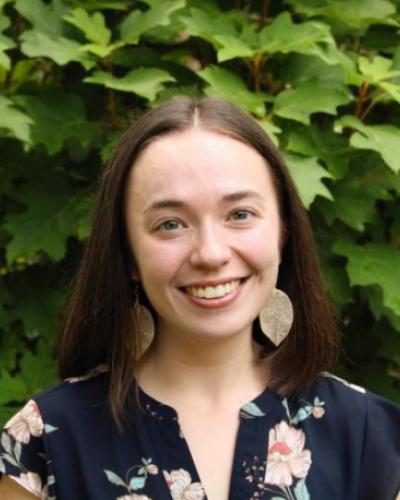 A person with shoulder‑length dark hair stands outdoors in front of dense green foliage. They are wearing a navy blouse with a floral pattern and large round earrings.